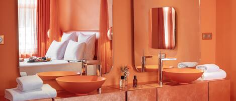 Un baño moderno con un espejo grande, dos lavamanos y una cama visible a través de la ventana.