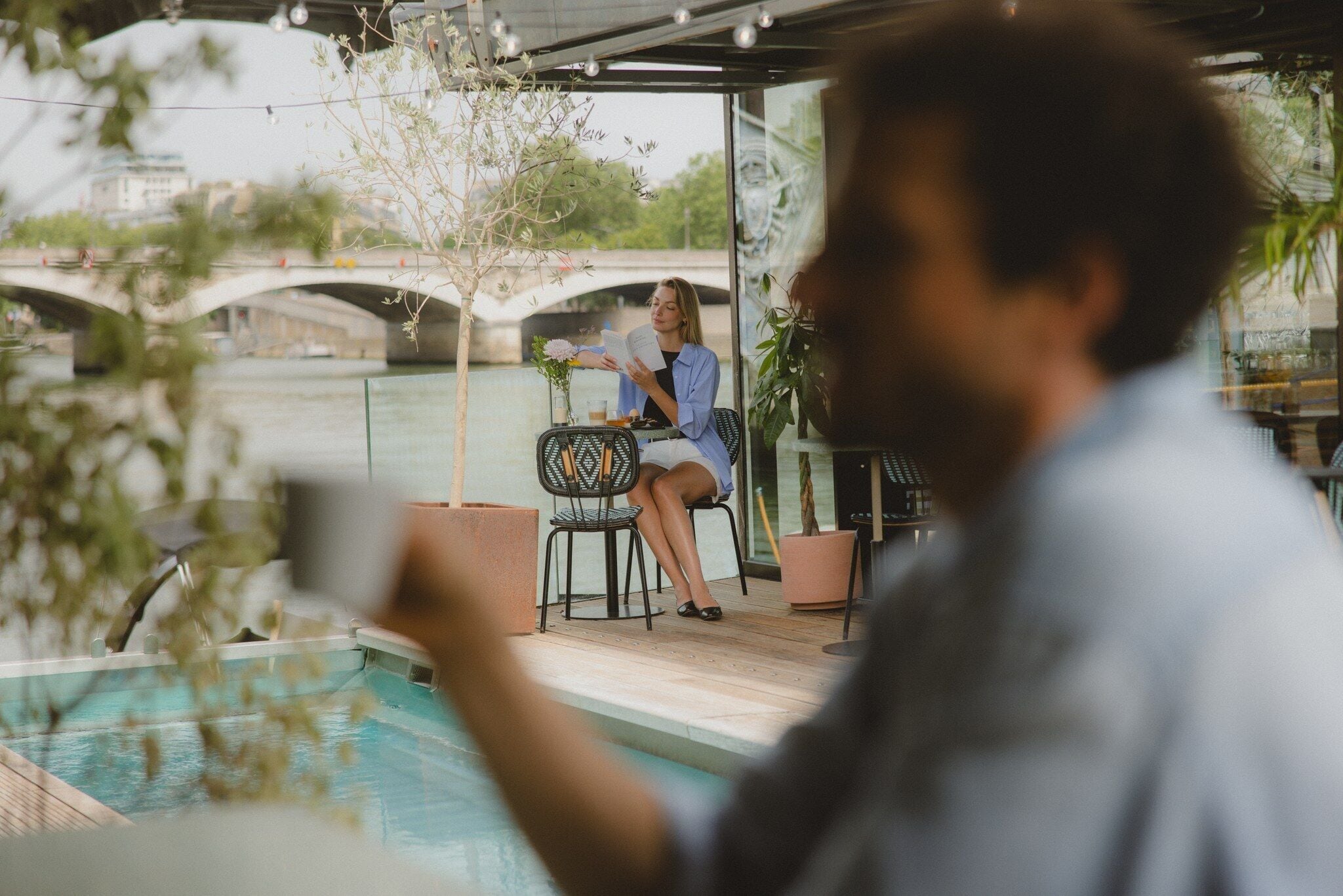 A person reading a book at a table by a poolside.