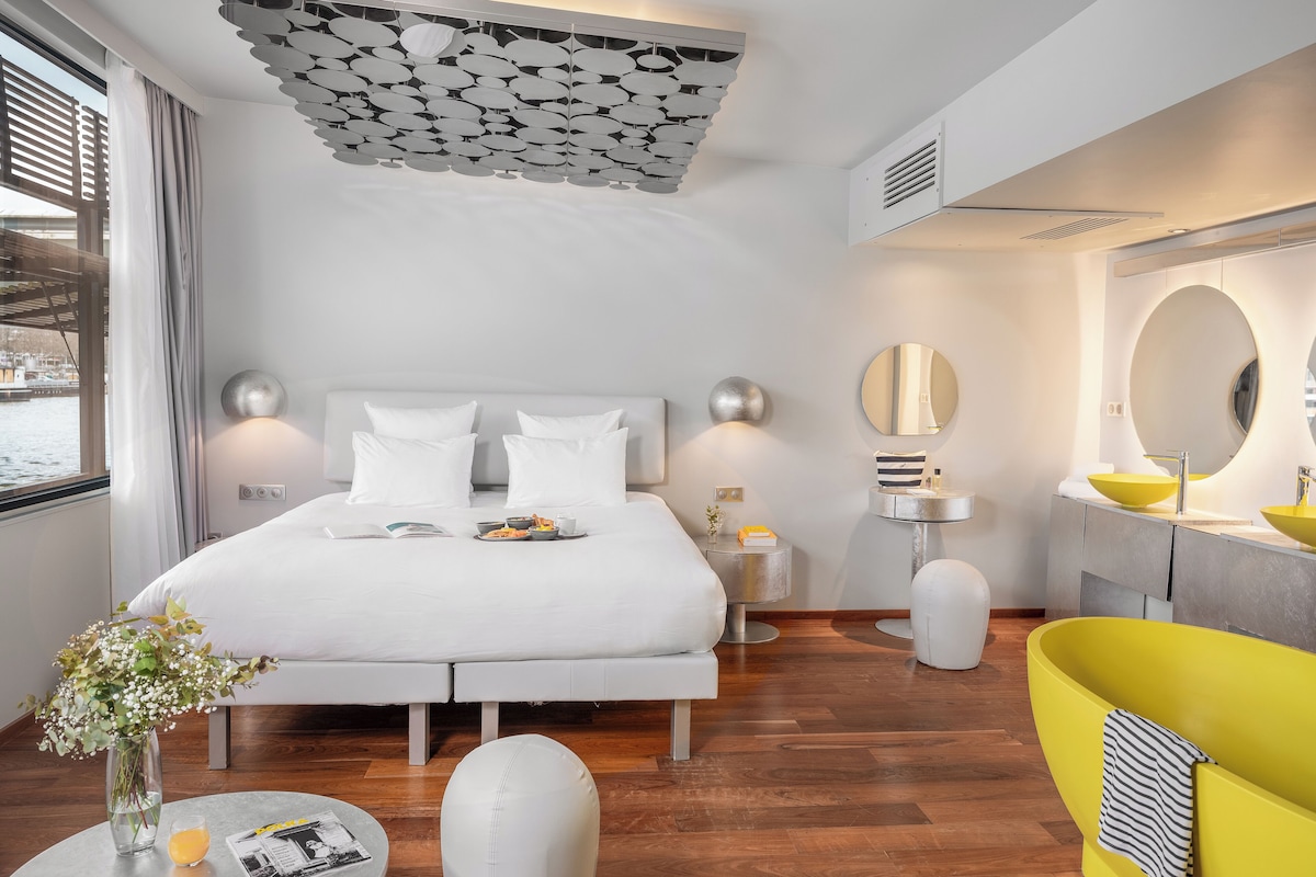 A modern hotel room with a large bed, a yellow bathtub, a round mirror, and a view of the outdoors.