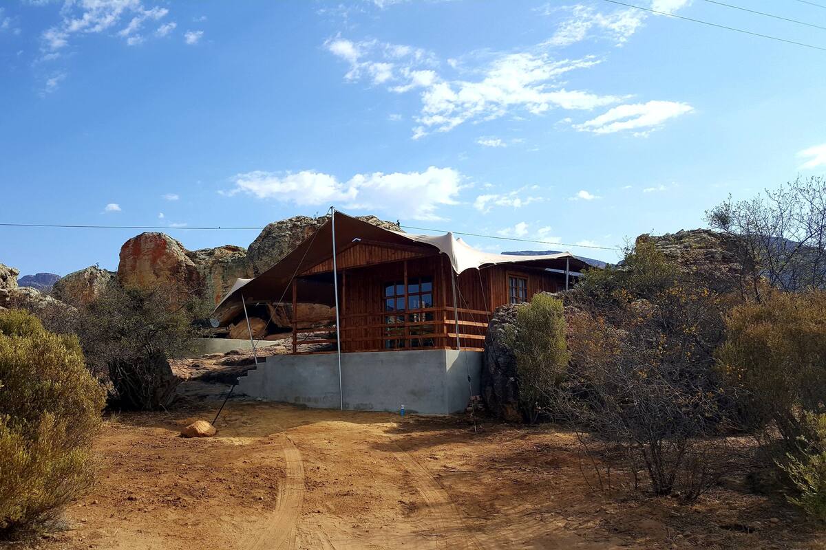 A rustic wooden cabin with a large rock formation in the background, surrounded by sparse vegetation.