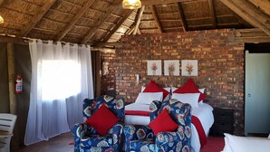 A room with a brick wall, a bed with white and red bedding, two patterned chairs, a white curtain, and a ceiling with wooden beams. - de Pakhuys (Cederberg)