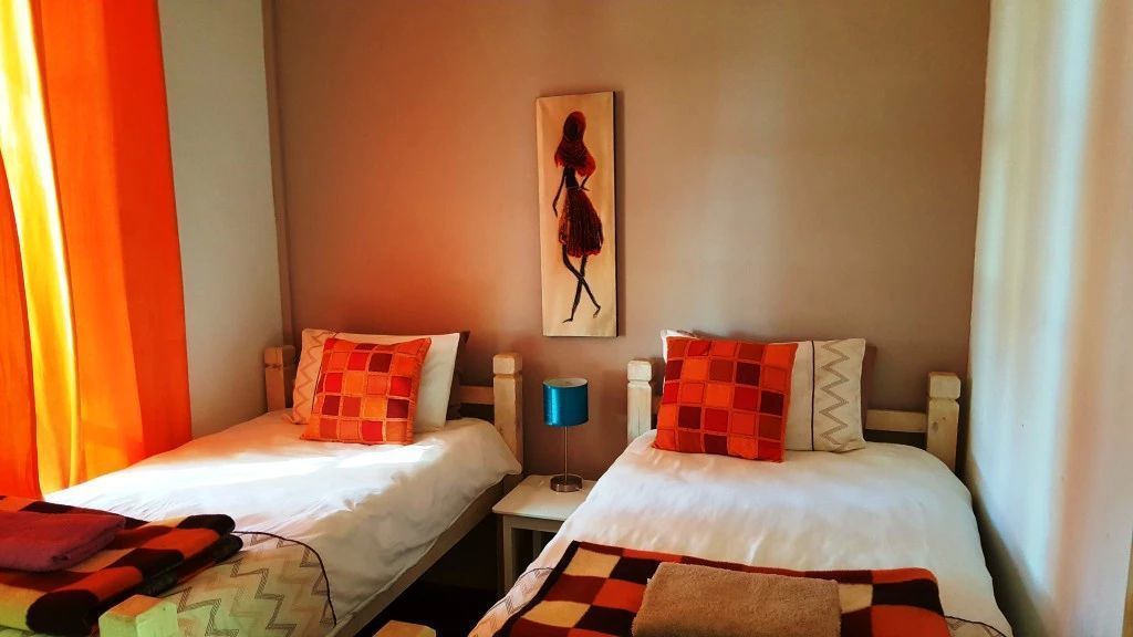 Dos camas individuales con almohadas de cuadros rojos y blancos, una lámpara de noche azul y un cuadro en la pared.