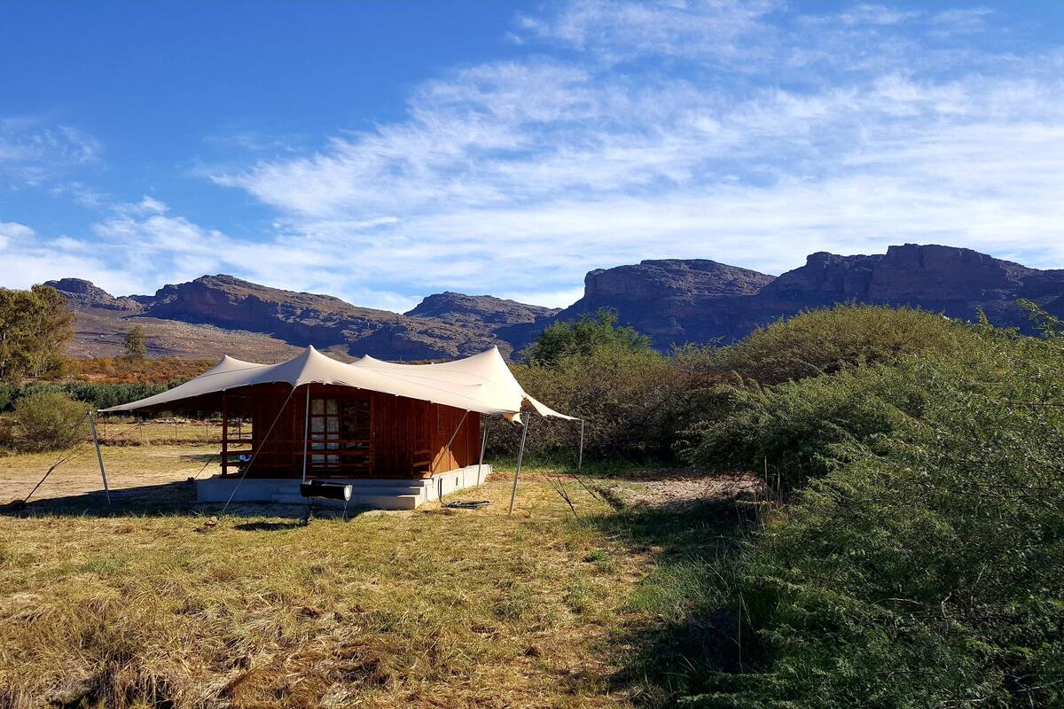 A wooden cabin with a large canopy, surrounded by dry grass and bushes, with mountains in the background.