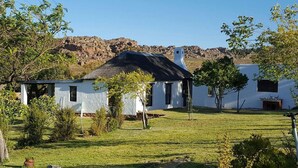 A white building with a black roof, surrounded by a grassy area and trees, with a rocky hill in the background. - de Pakhuys (Cederberg)