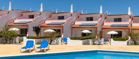 A swimming pool with blue water, surrounded by white buildings with orange roofs and multiple chimneys. There are blue lounge chairs and umbrellas near the pool.