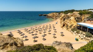 Une plage dotée de nombreux parasols et de chaises longues, un littoral rocheux et un bâtiment surmonté d’un toit bleu.