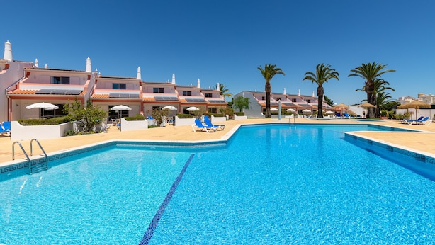 Une piscine entourée de bâtiments dotés de balcons et de palmiers, avec des chaises longues et des parasols à proximité.