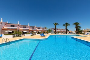 Una piscina con sillones de descanso y sombrillas, rodeada de edificios con balcones y palmeras.