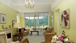 A spacious living room with a fireplace, comfortable seating, a dining table with a vase of flowers, and large windows offering a view of the outdoors. - Plas Maenan Country House (Llanrwst)