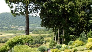 A lush garden with various trees and shrubs, a well-maintained lawn, and a distant view of a town. - Plas Maenan Country House (Llanrwst)