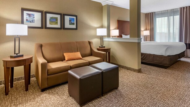 A hotel room with a brown sofa, a small ottoman, a bed, and two bedside tables with lamps. There are framed pictures on the wall. - Comfort Inn PA Turnpike - 1-81 (Carlisle)
