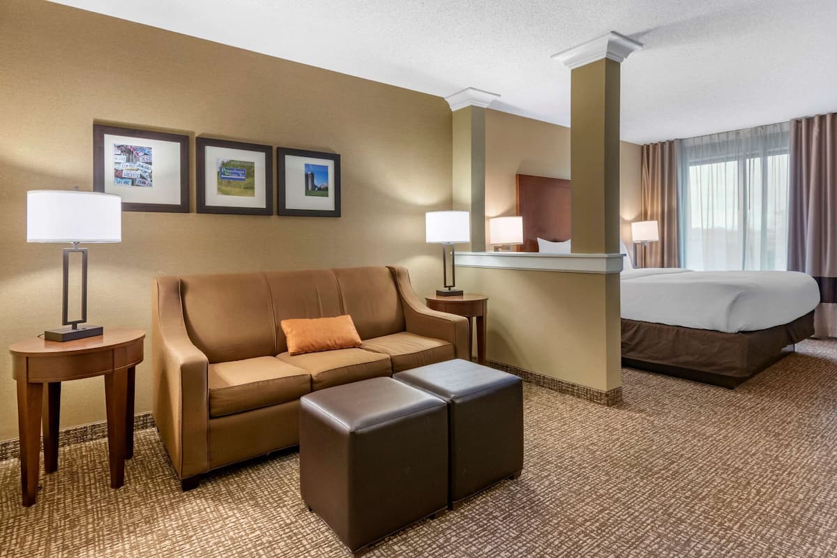 A hotel room with a brown sofa, a small ottoman, a bed, and two bedside tables with lamps. There are framed pictures on the wall.