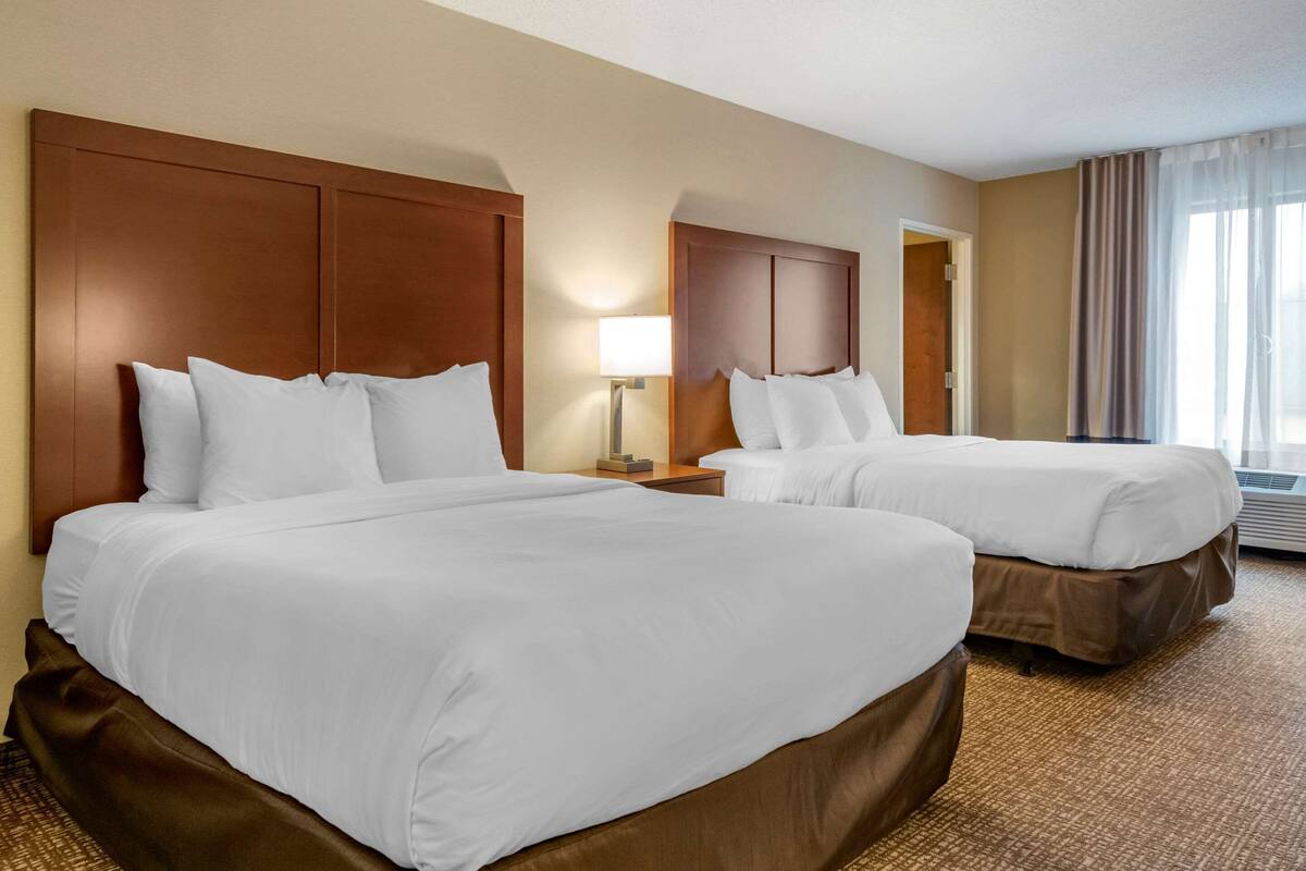 A hotel room with two beds, a nightstand with a lamp, and a large window with curtains.