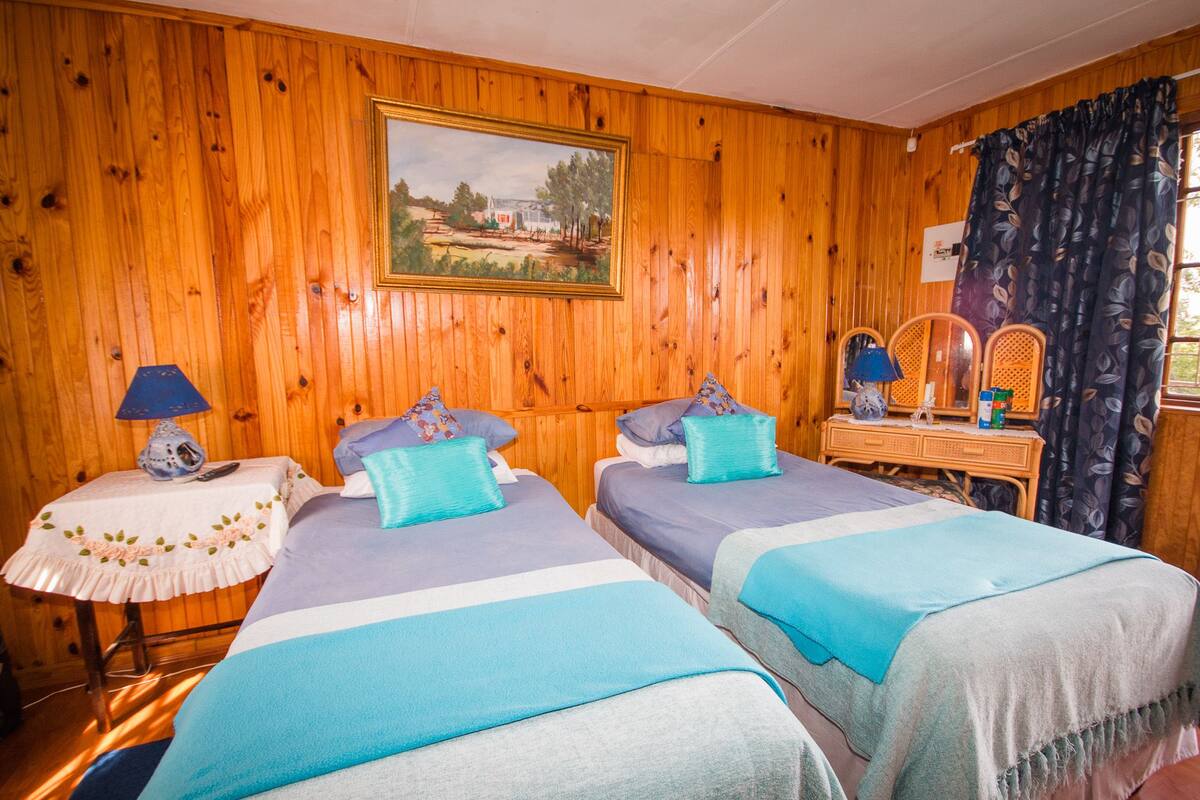 A room with two beds, wooden walls, a painting, a dresser with mirrors, and a window with curtains.