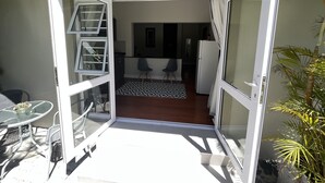 An open sliding door leading to a patio with a dining table and chairs, a kitchen area with a refrigerator, and a patterned rug.