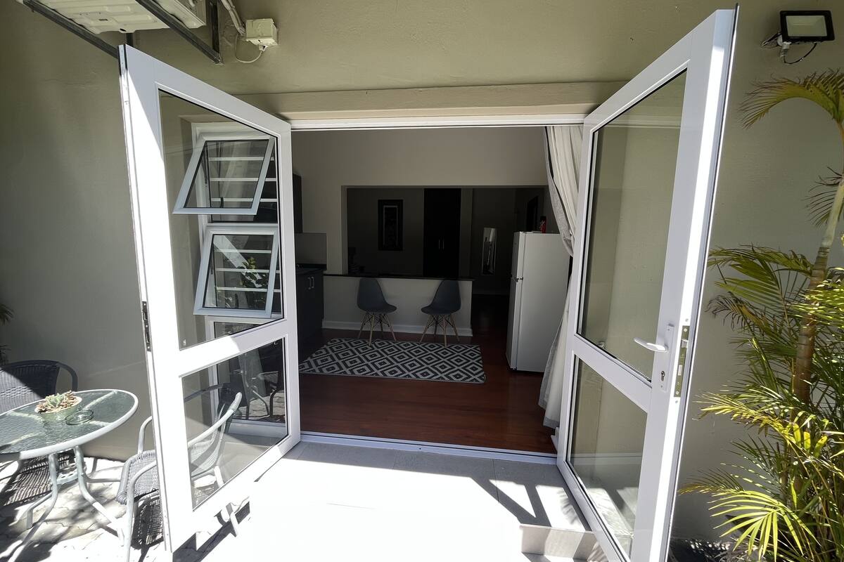 An open sliding door leading to a patio with a dining table and chairs, a kitchen area with a refrigerator, and a patterned rug.