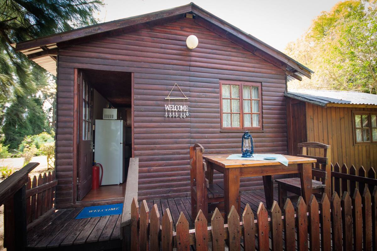 A wooden cabin with a 'WELCOME' sign, a table with a lantern, and a wooden fence.