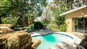 A swimming pool with a rock formation, surrounded by trees and a building with a glass door. - Homestead B & B (Addo)