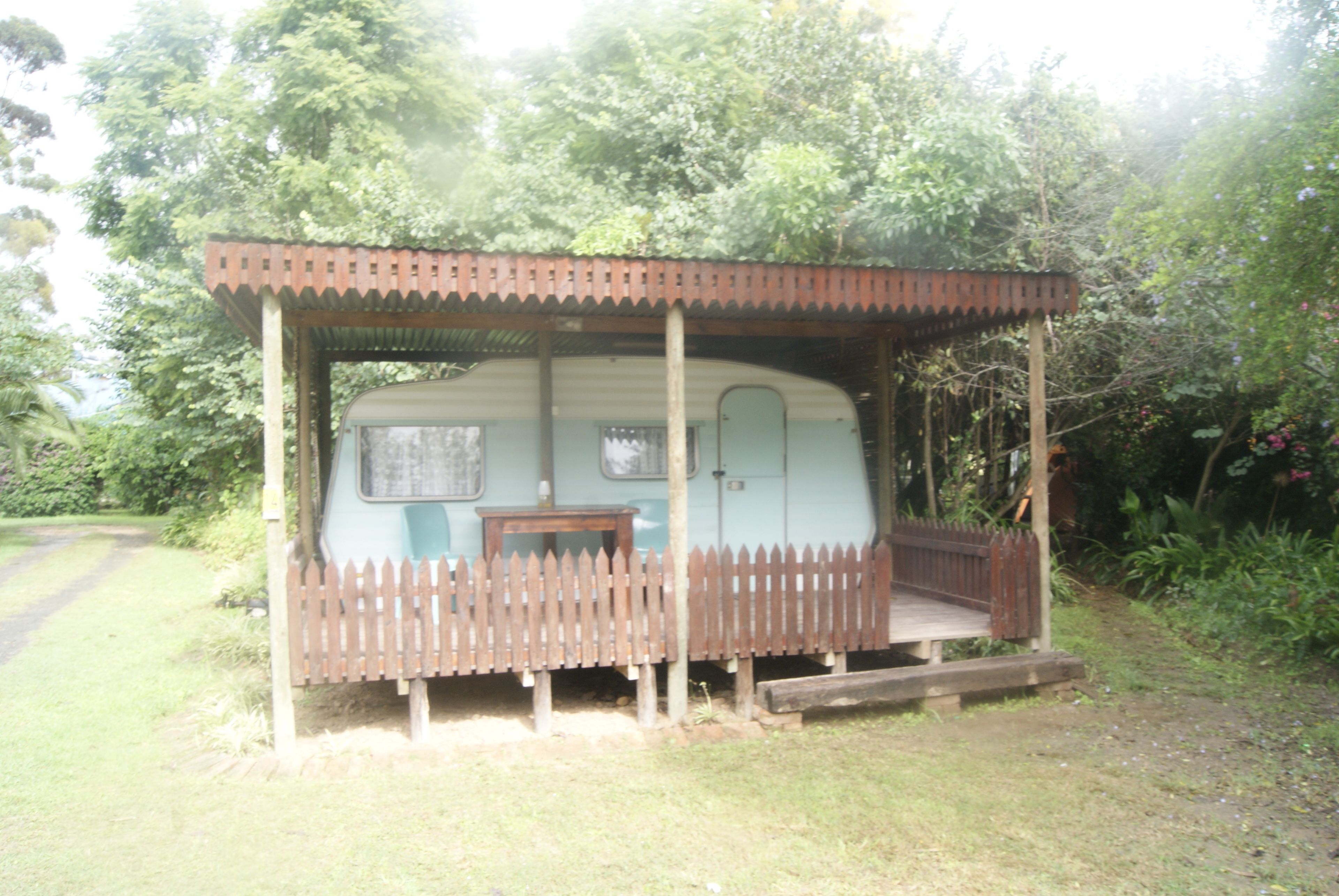 A small, single-story RV with a porch and a table, surrounded by lush greenery.