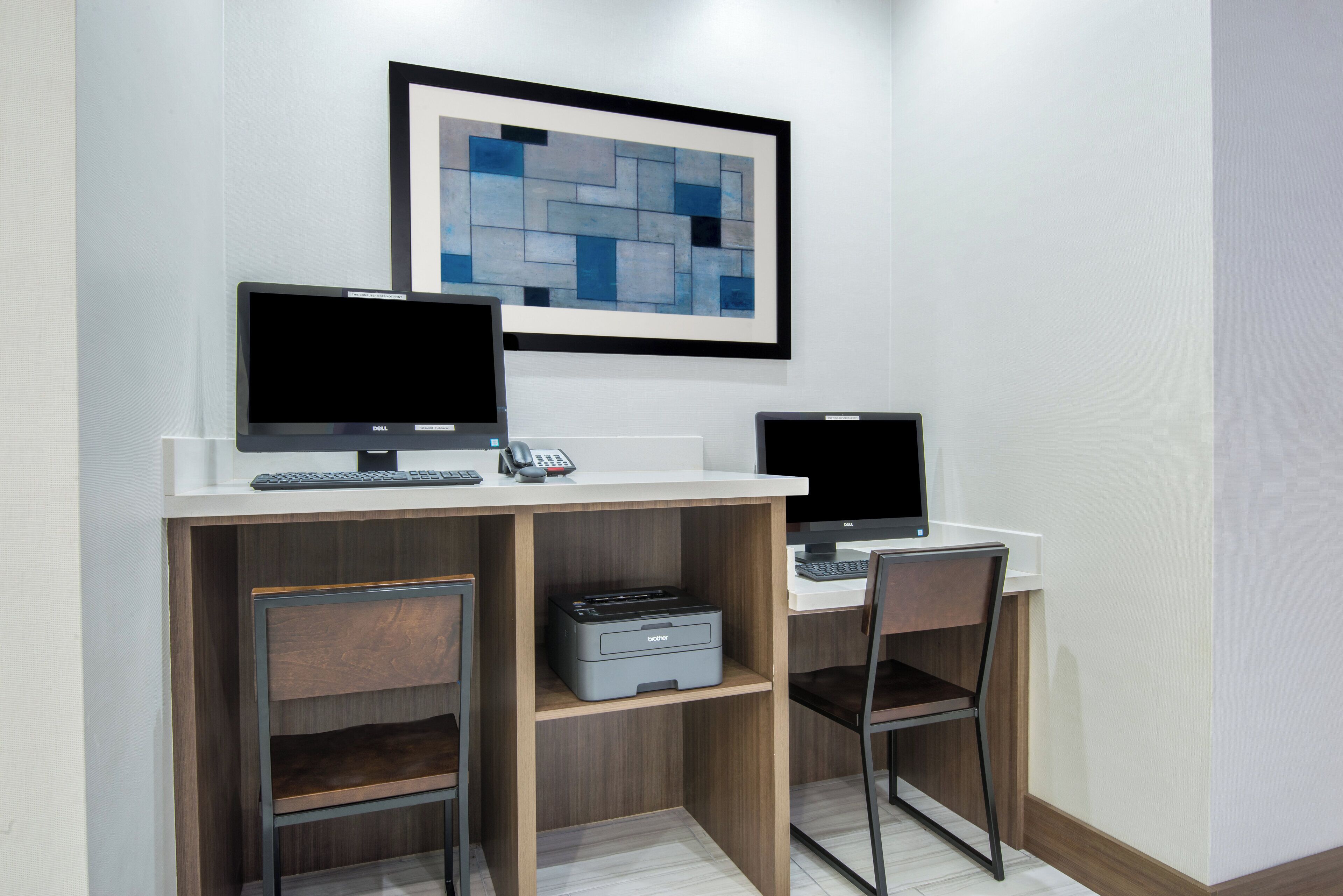 A modern office space with two desks, each with a computer monitor, keyboard, and mouse. There are two chairs, one at each desk. A printer is placed on the floor between the desks. A framed abstract painting hangs on the wall above the desks.