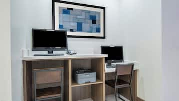 A modern office space with two desks, each with a computer monitor, keyboard, and mouse. There are two chairs, one at each desk. A printer is placed on the floor between the desks. A framed abstract painting hangs on the wall above the desks.