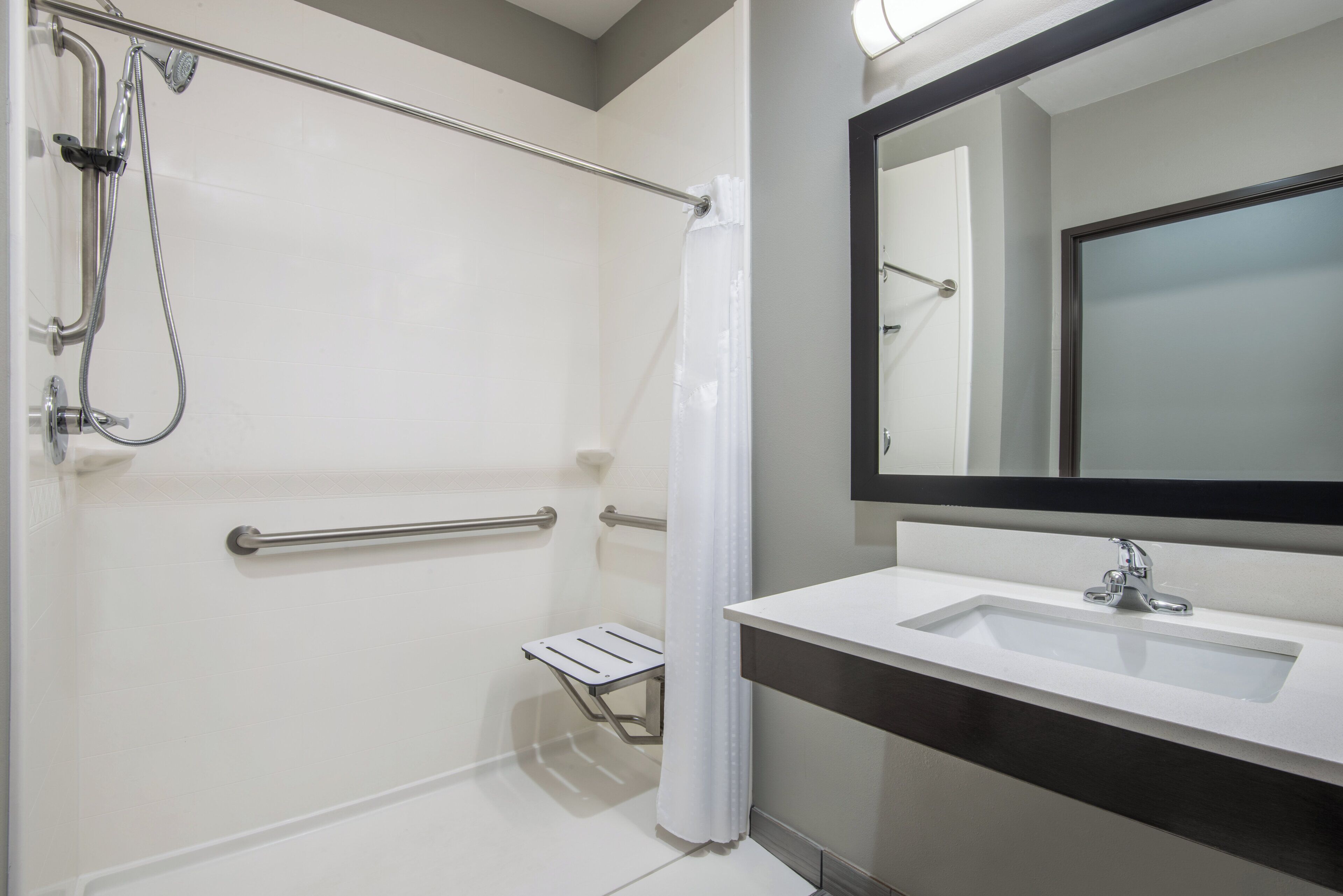 A modern bathroom with a walk-in shower, grab bars, a small step stool, and a large mirror.