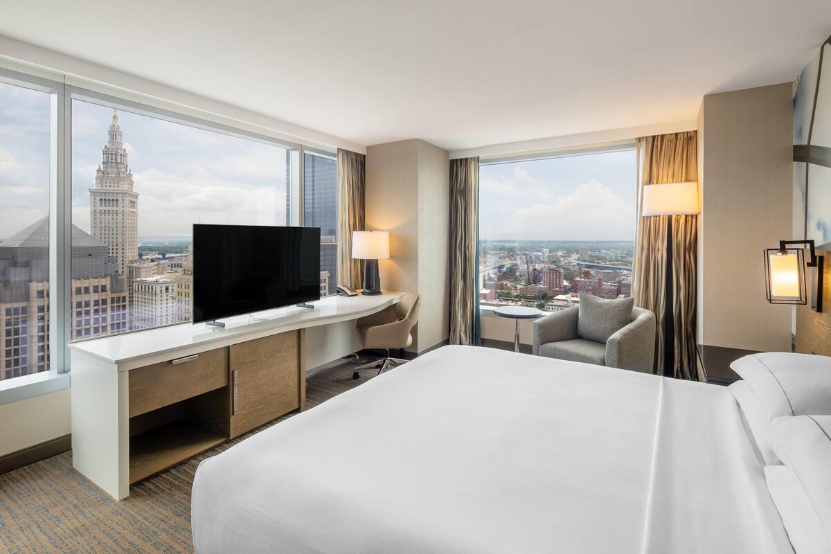 A hotel room with a large bed, a desk with a TV, a chair, a small table, and a view of a cityscape.