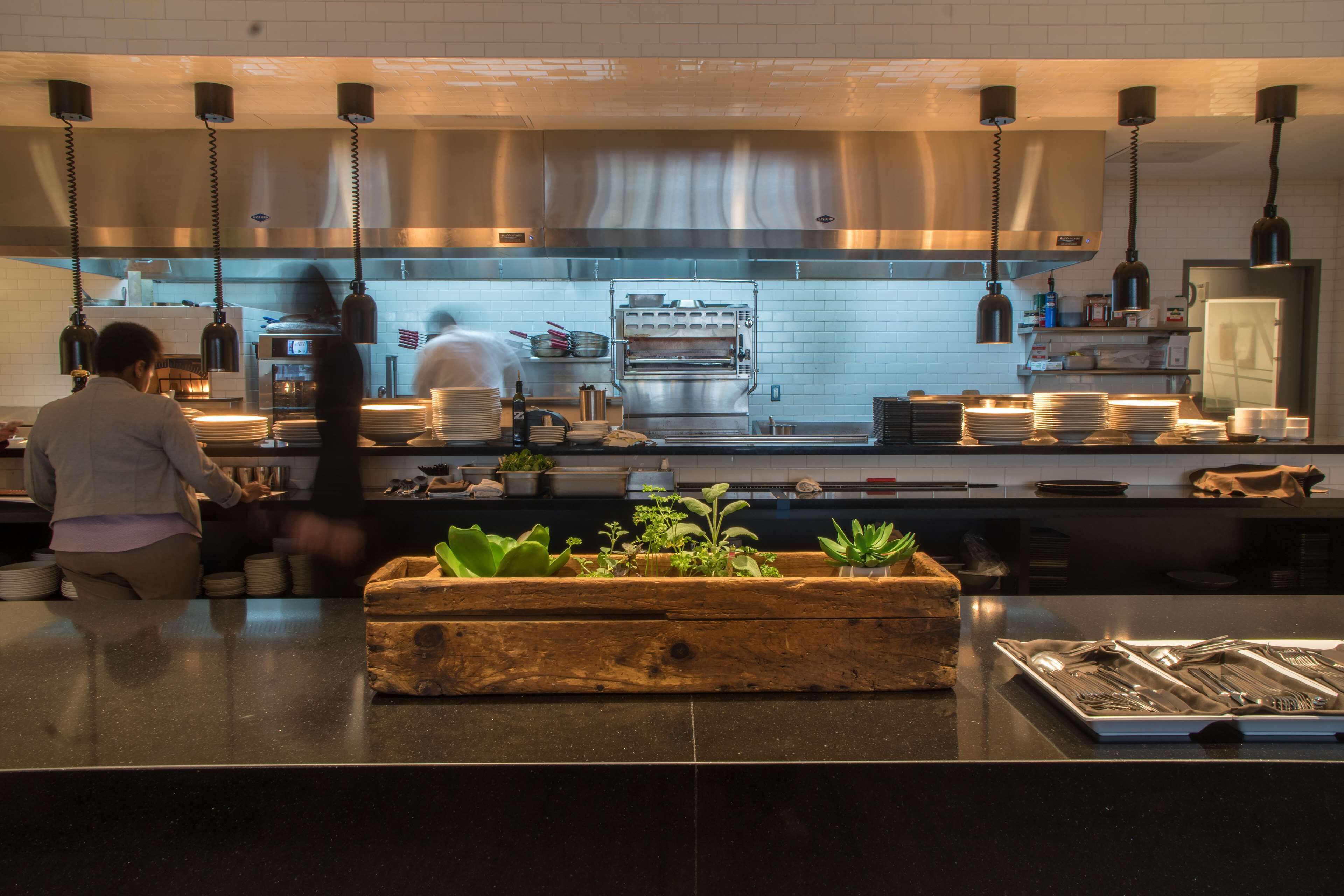 A modern kitchen with stainless steel appliances, hanging pendant lights, and a wooden planter with green plants on the counter.