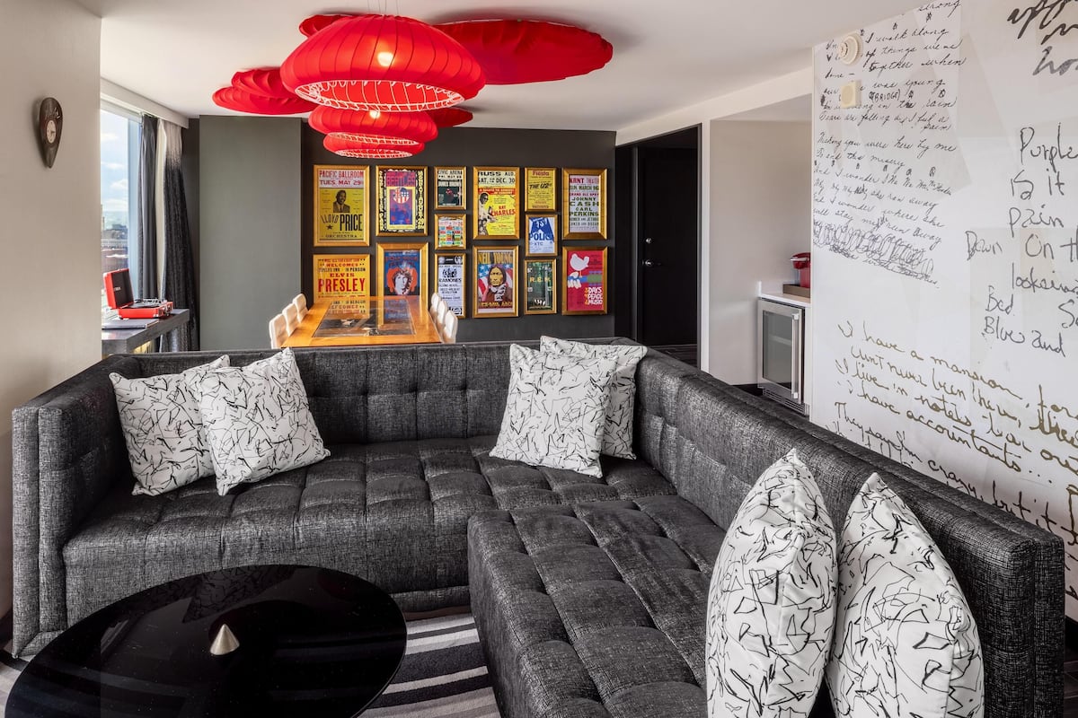A modern living room with a sectional sofa, a black coffee table, and red hanging lamps. There are framed posters on the wall and a whiteboard with handwritten notes.