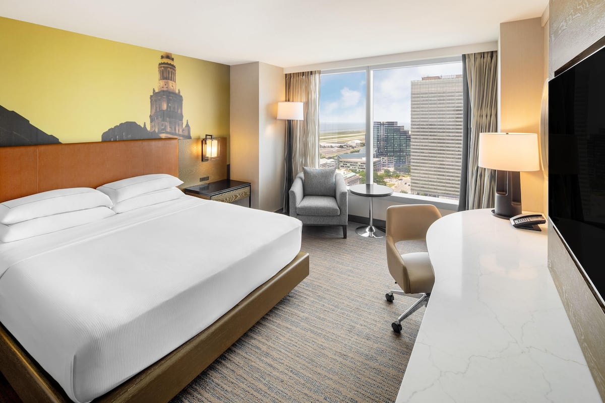 A hotel room with a large bed, a desk with a chair, a TV, a lamp, and a view of a cityscape.