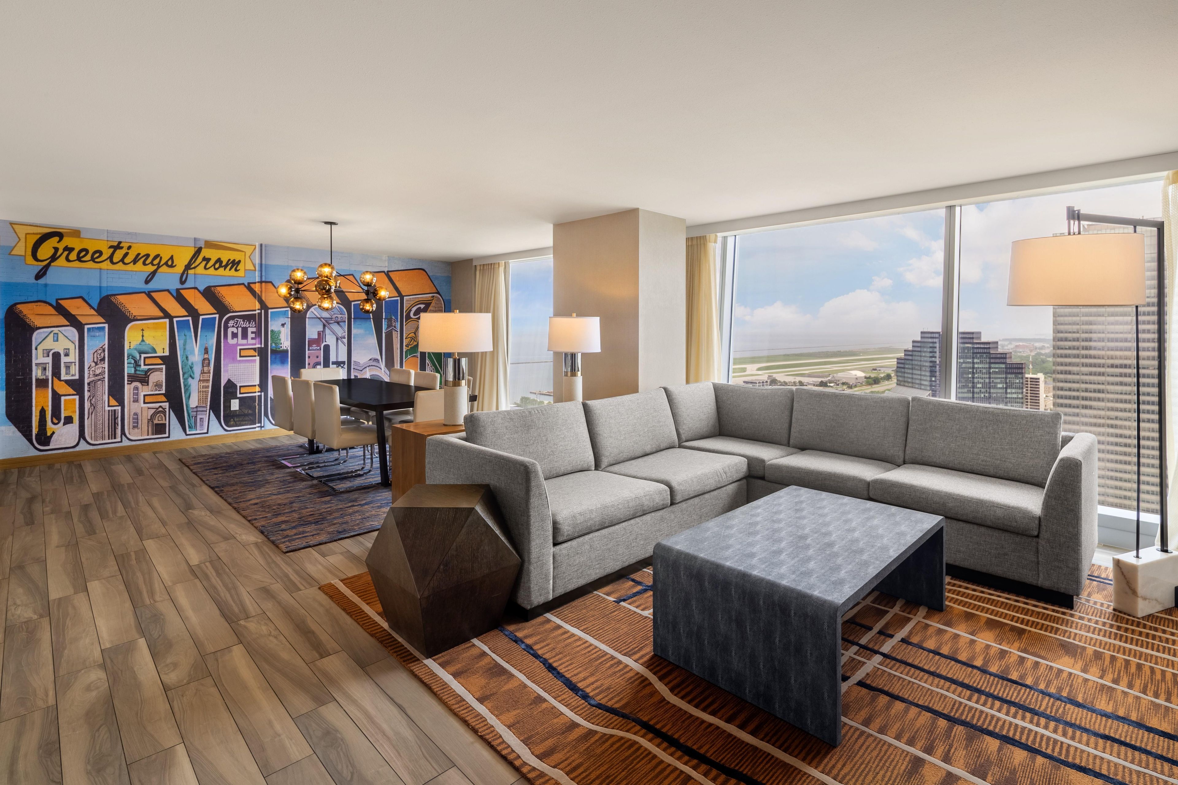 Ein modernes Hotelzimmer mit einer großen, grauen Eckcouch, einem kleinen Couchtisch und Blick auf die Stadt durch große Fenster.