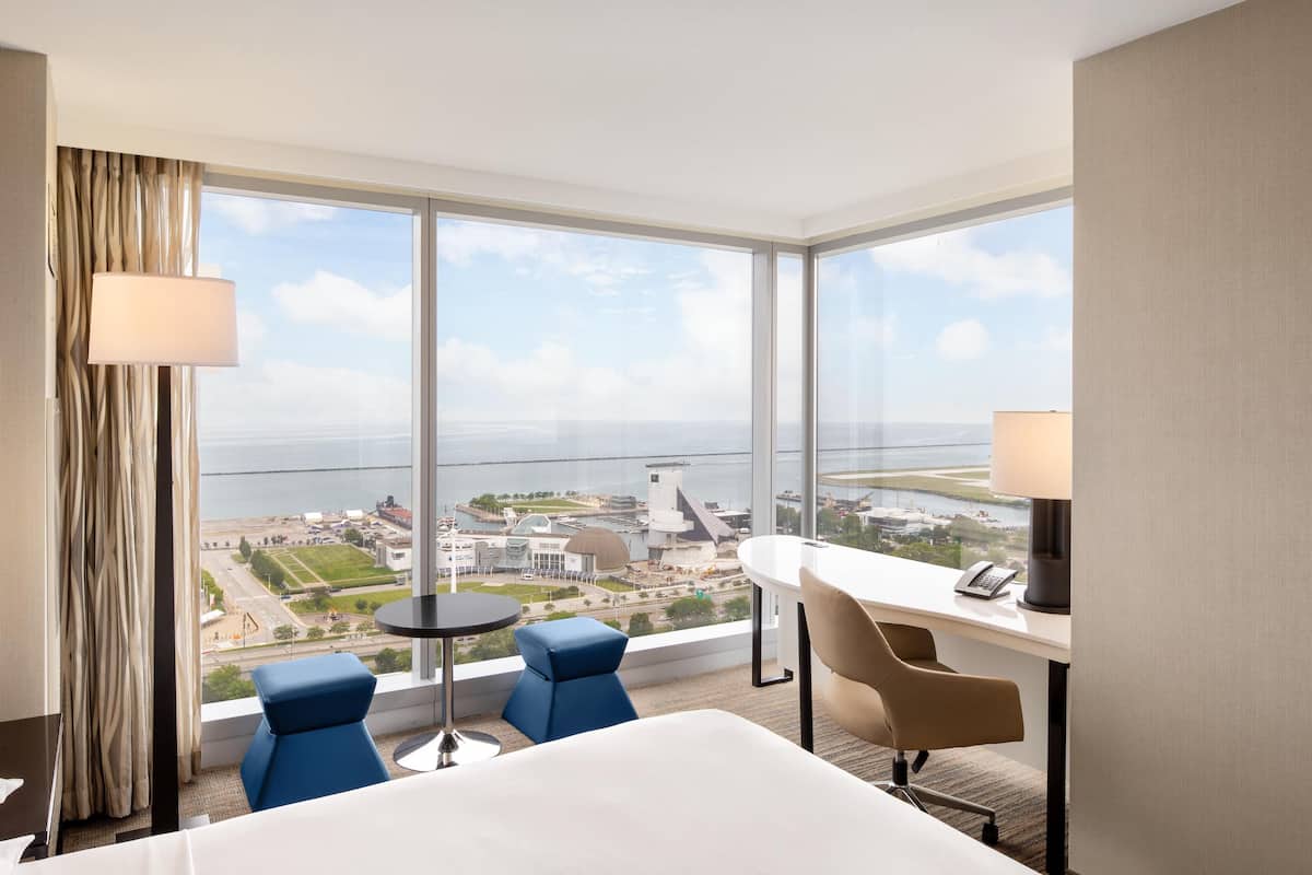 A hotel room with a large window offering a view of the city and the sea. There is a bed, two chairs, a desk, and a lamp.