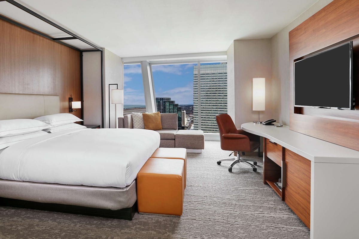 A hotel room with a bed, a desk, a chair, and a large window with a city view.