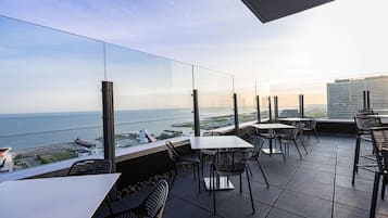 A terrace with tables and chairs, overlooking a body of water and a distant cityscape.