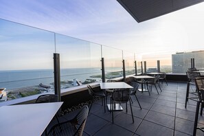 Een terras met tafels en stoelen, met uitzicht op een waterpartij en een verafgelegen stadsbeeld.