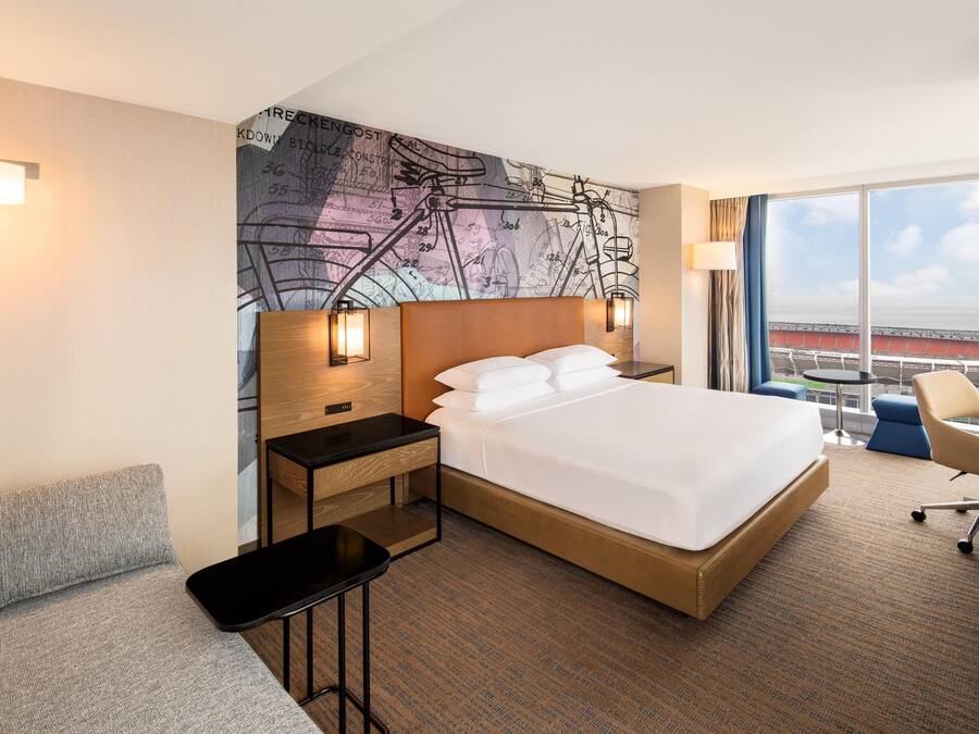 Habitación de hotel con una cama grande, un escritorio, una silla, una mesita y un amplio ventanal con vistas a un estadio.