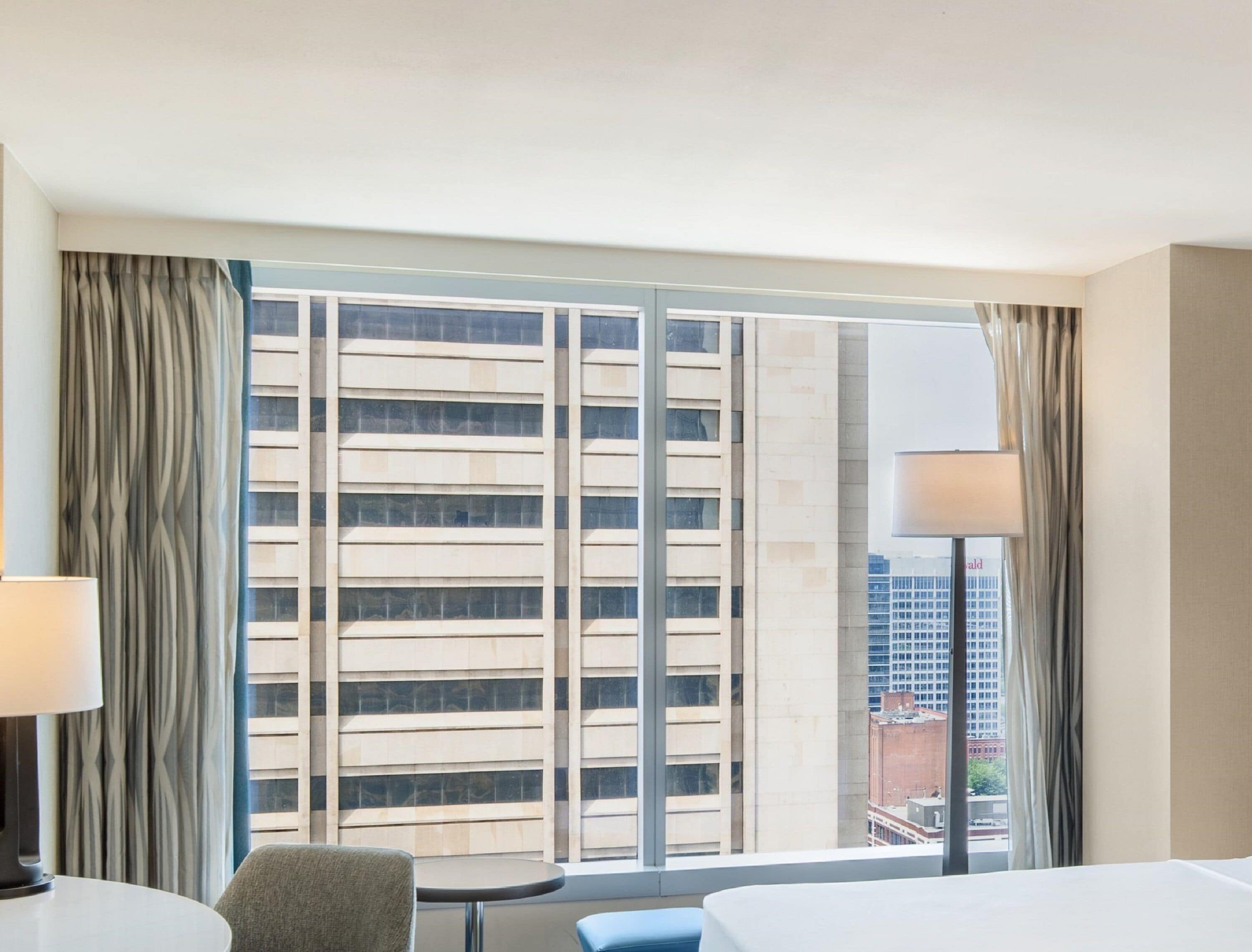 Une chambre d’hôtel avec une grande fenêtre, un lit, une chaise, une petite table et une lampe.