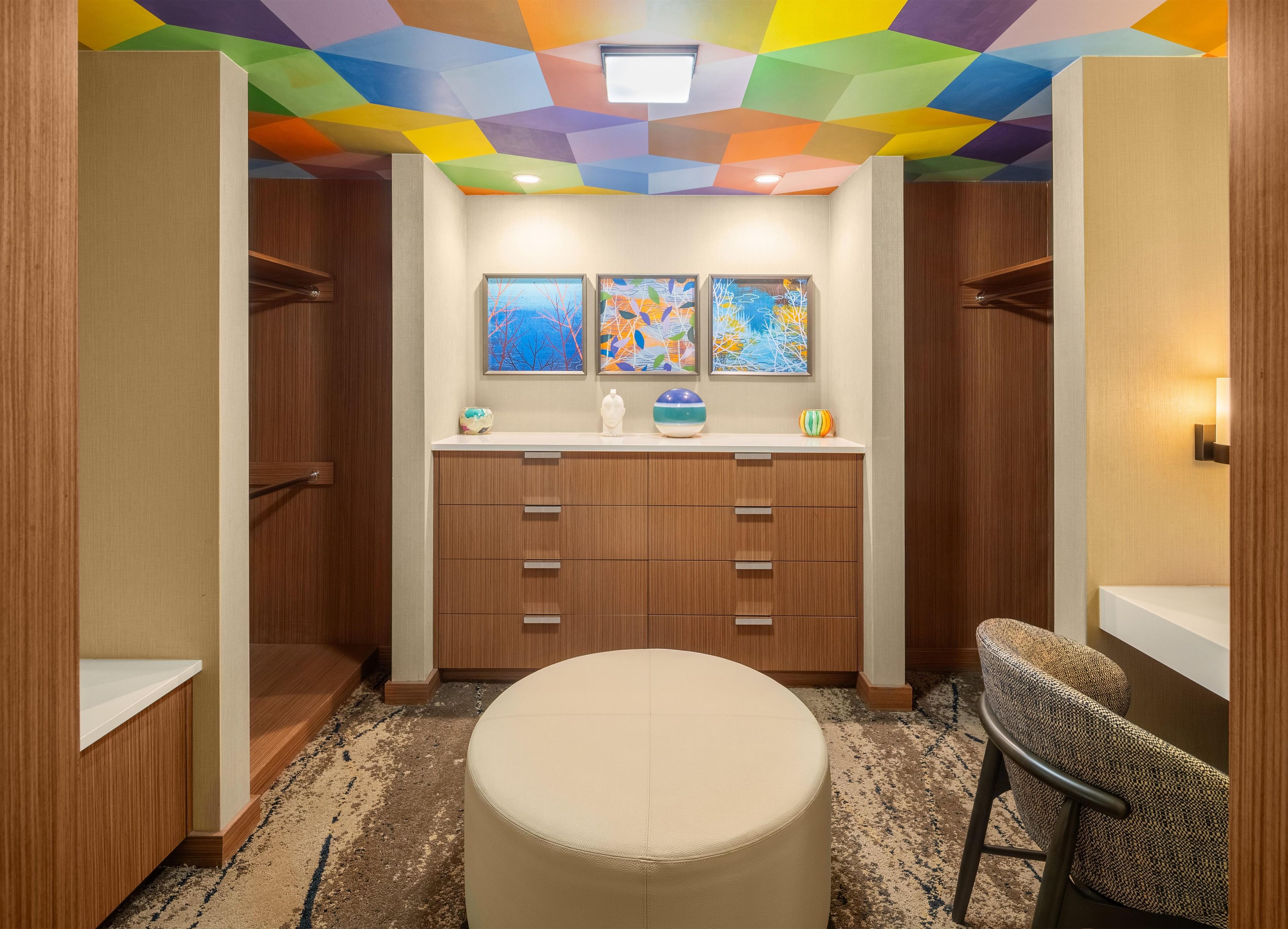 Une chambre d’hôtel avec un plafond orné de motifs géométriques, une armoire en bois, un pouf rond de couleur beige et une chaise à motifs.