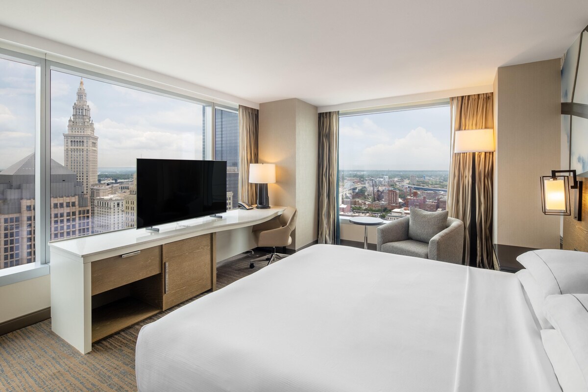 Ein Hotelzimmer mit einem großen Bett, einem Schreibtisch mit Fernseher, einem Sessel, einem kleinen Tisch und Blick auf die Stadt.