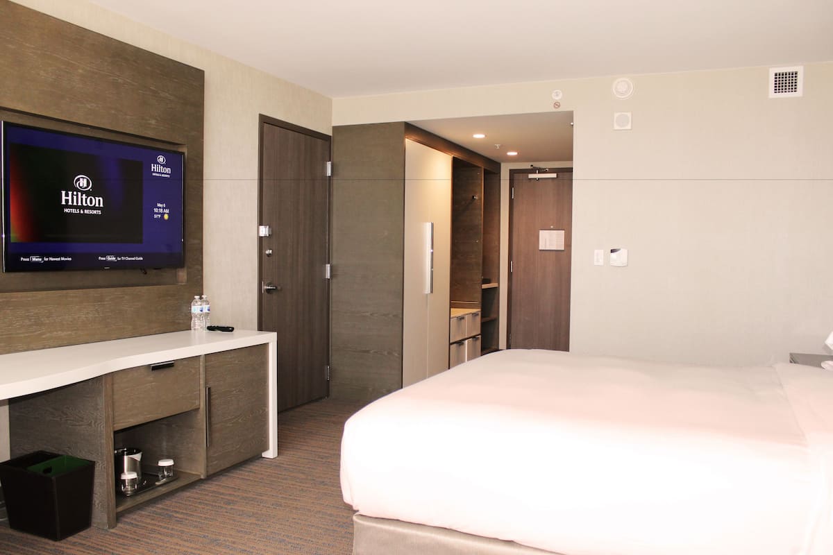 A modern hotel room with a flat-screen TV displaying the Hilton logo, a white desk with a bottle of water, a bed with a white comforter, and a walk-in closet.