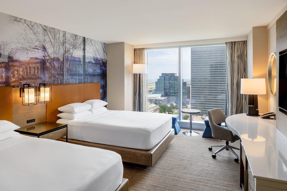 A hotel room with two beds, a desk, a chair, a large window with a city view, and a painting on the wall.