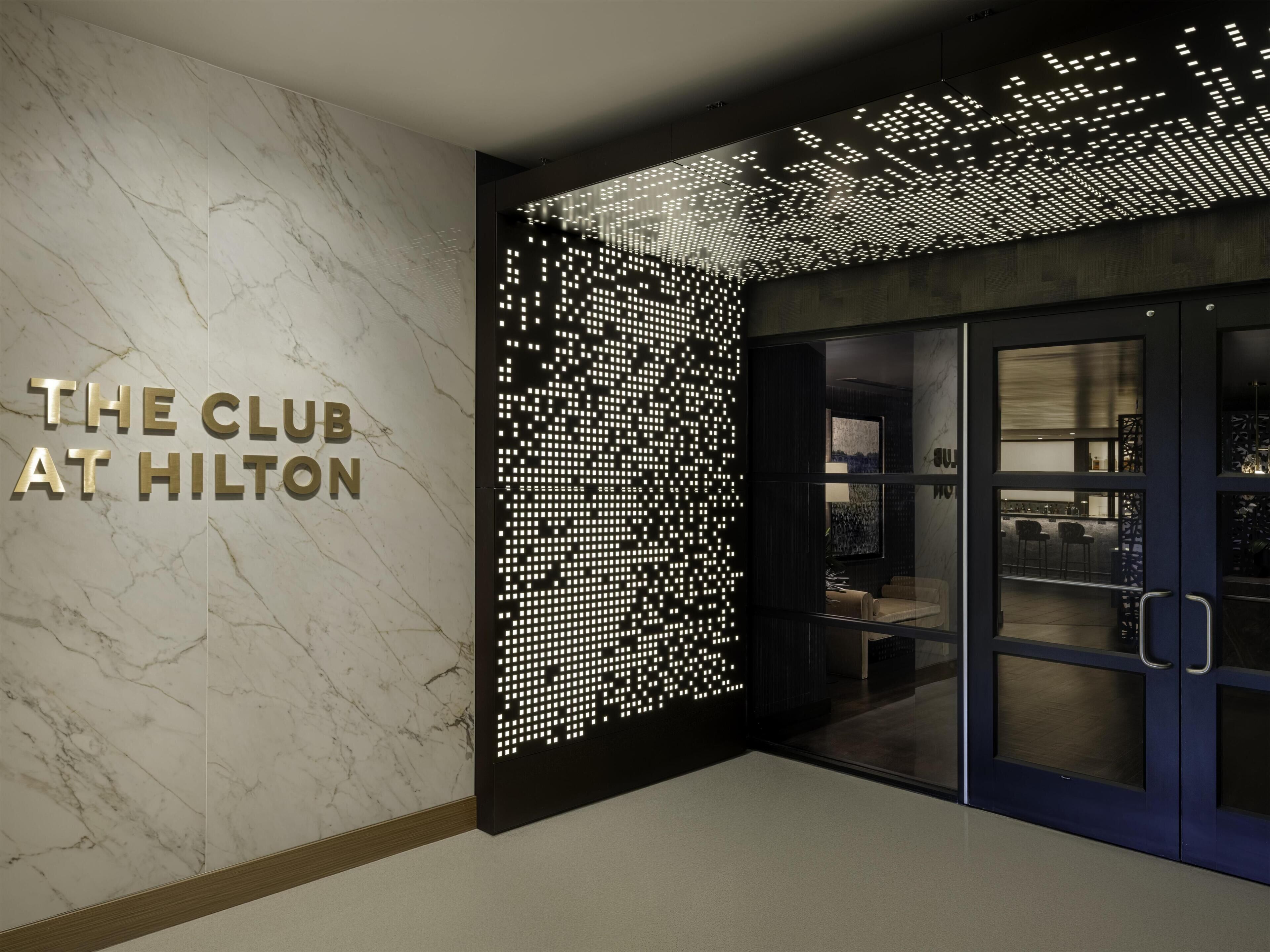 Una entrada de hotel moderna con una pared de mármol, un letrero que dice 'THE CLUB AT HILTON' y un panel luminoso decorativo.