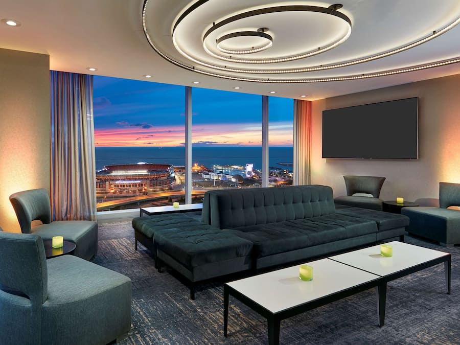 A modern hotel room with a large window offering a city view, a comfortable seating area with a sofa and chairs, a flat-screen TV, and ambient lighting.