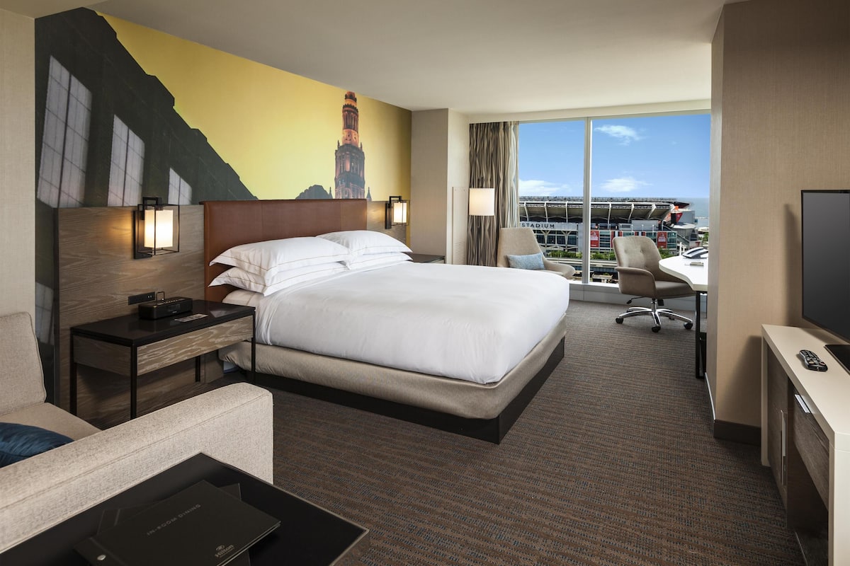 A hotel room with a large bed, a desk, a chair, and a view of a stadium.