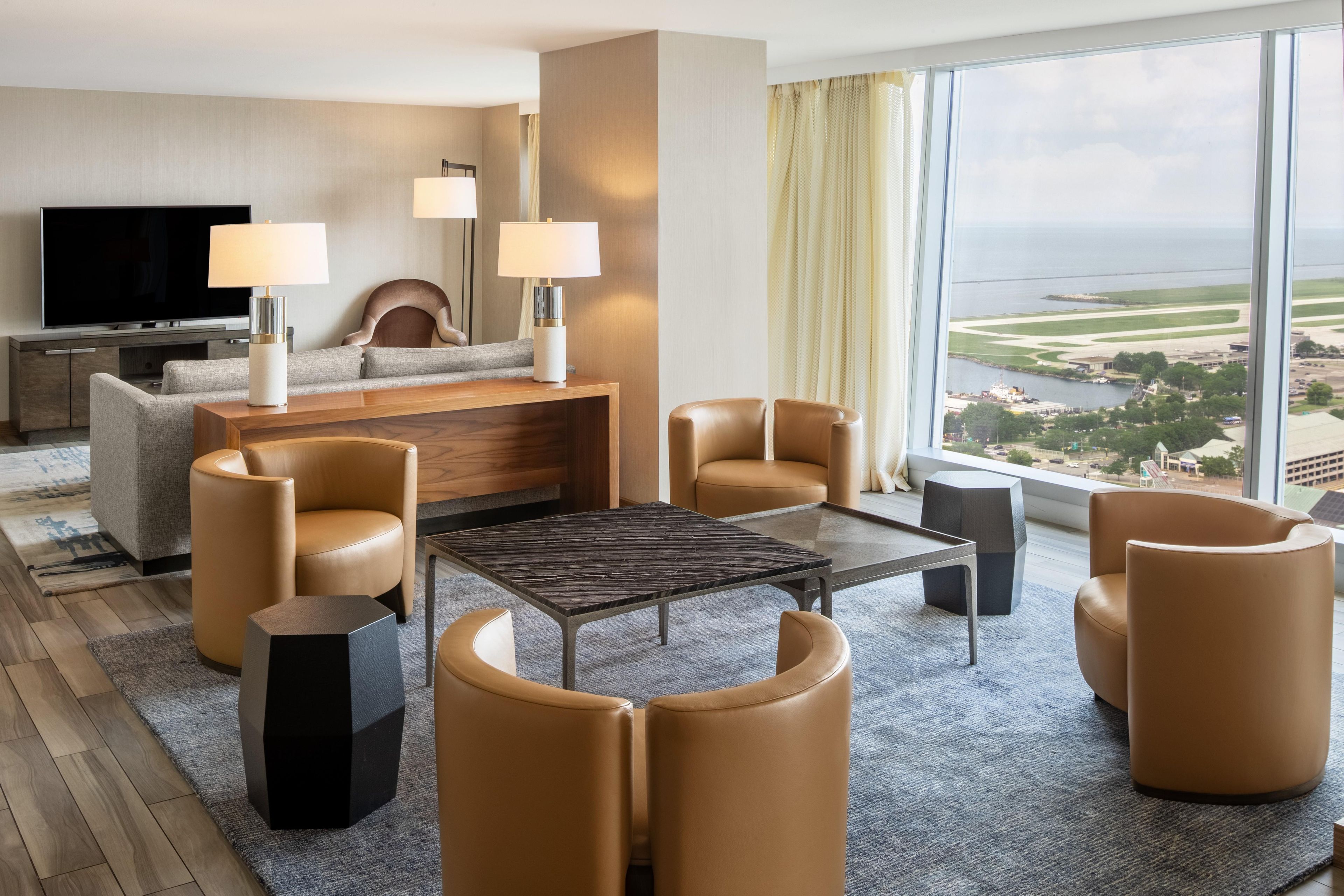 A modern hotel room with a large window offering a view of the coastline, a flat-screen TV, a sofa, two armchairs, a coffee table, and a side table with lamps.