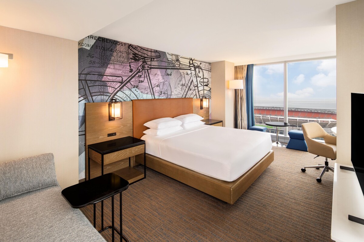 Ein Hotelzimmer mit einem großen Bett, einem Schreibtisch, einem Sessel, einem kleinen Tisch und einem großen Fenster mit Blick auf ein Stadion.