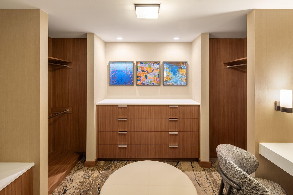 Ein Hotelzimmer mit einem Holzschrank, einer weißen Kommode, einem runden Spiegel und drei gerahmten Kunstwerken an der Wand.