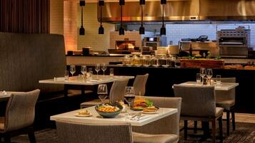Een restaurant met meerdere tafels en stoelen, een keuken op de achtergrond, en diverse gerechten en drankjes op de tafels.