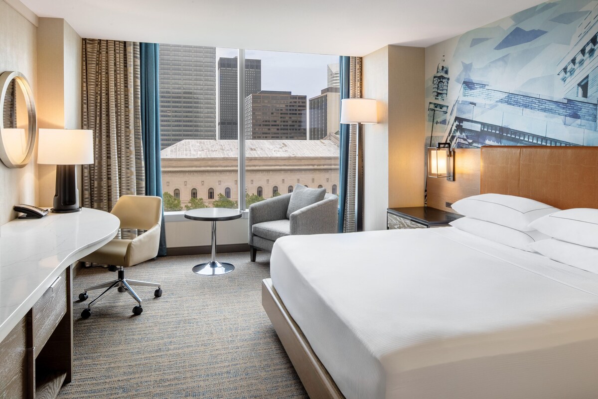Ein Hotelzimmer mit einem großen Bett, einem Schreibtisch, einem Sessel, einem kleinen Tisch und Blick auf Stadtgebäude durch das Fenster.