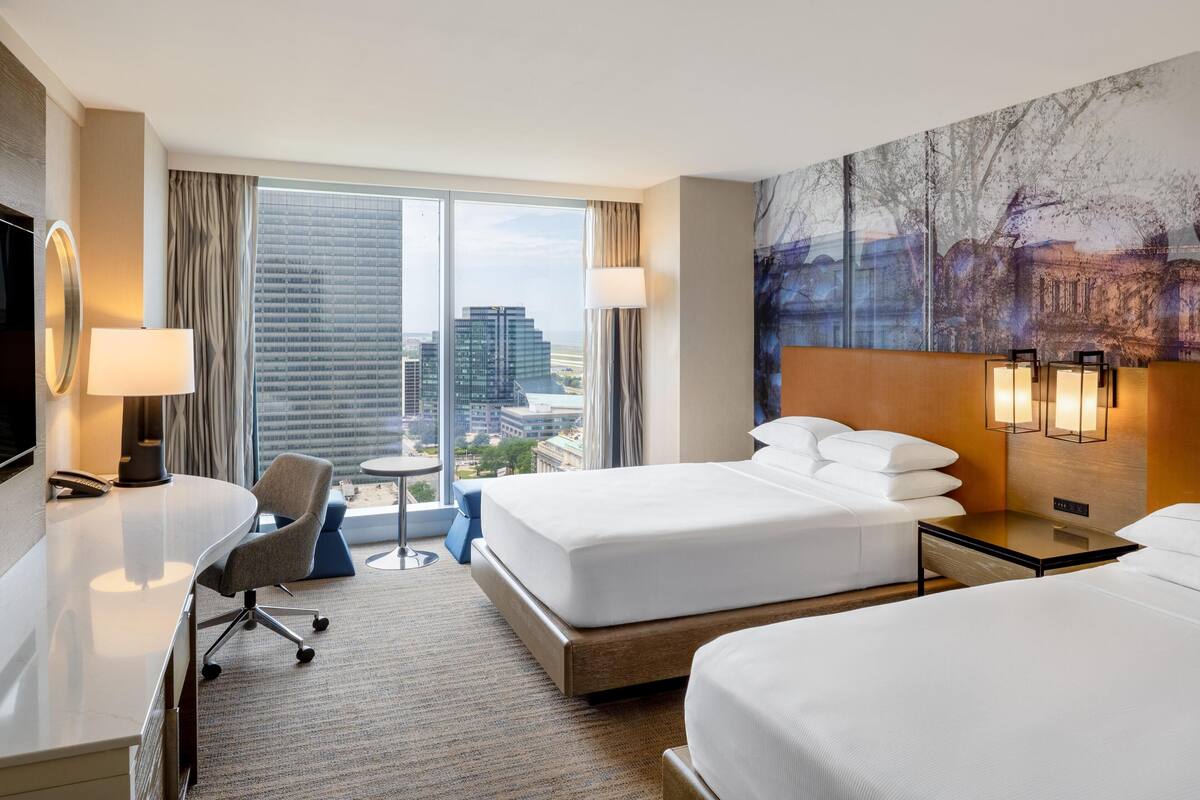 A hotel room with a desk, two beds, a chair, a large window with a city view, and a painting on the wall.
